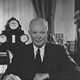 President Eisenhower behind his desk in the Oval Office, holding some papers, with his eyeglasses resting in front of him.