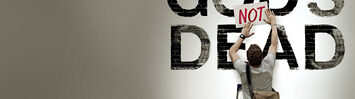 A man places a sign that reads 'NOT' in red, capital letters, on top of a wall that has the message 'God's Dead' painted on it to change the meaning of the sentence.