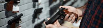 Various guns are displayed on a wall behind a man's hands holding a pistol. 