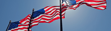 Three American flags blow in the wind against a blue, sunny sky. 
