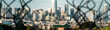 A highway and city skyline are seen through a hole in a chain link fence.
