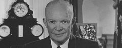 President Eisenhower behind his desk in the Oval Office, holding some papers, with his eyeglasses resting in front of him.